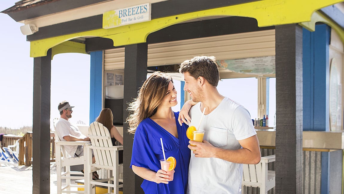 A couple is standing under a covered outdoor bar with a sign that reads Breezes Pool Bar & Grill.
