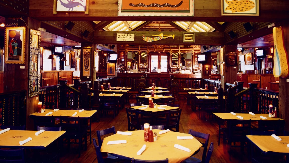 a dining room with wooden tables and chairs, yellow tablecloths, and a bar area in the background
