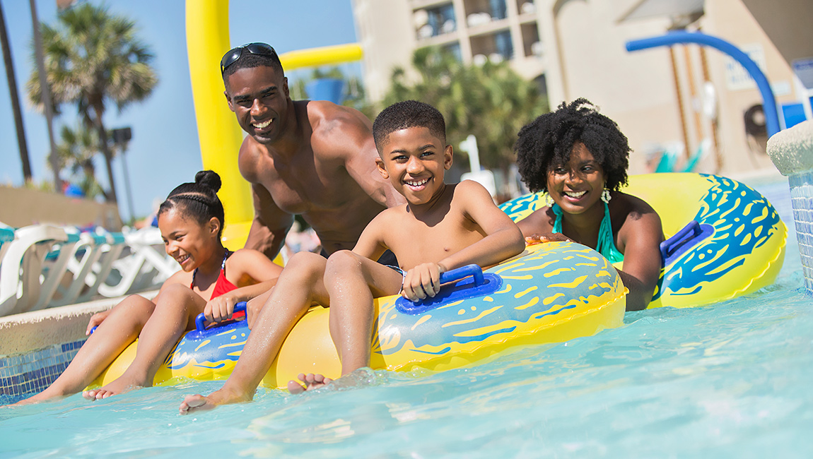 A family of four is enjoying a sunny day in the pool, floating on yellow and blue inflatable tubes.