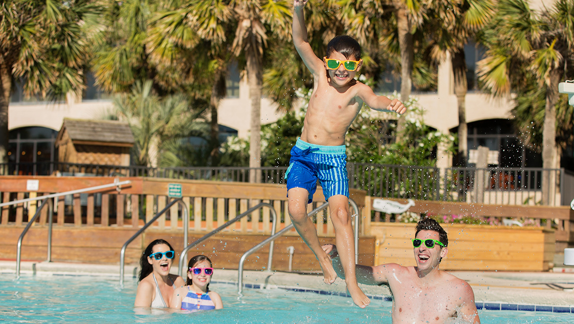 A family enjoys a sunny day at the pool, with a boy jumping in and others swimming and laughing.