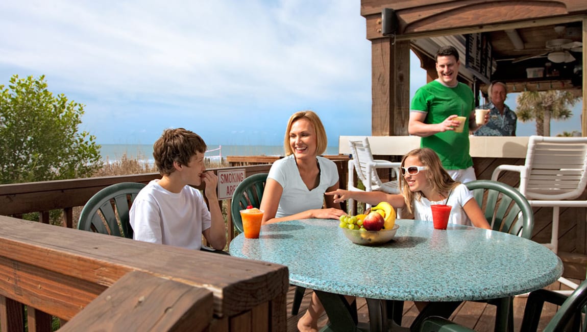 A family of four enjoys a meal at an outdoor table with a fruit bowl and drinks.