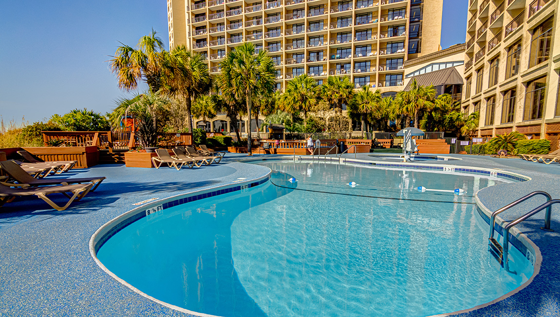 A curved pool with lounge chairs and palm trees in front of a hotel building.